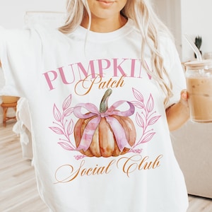 May include: White t-shirt with a watercolor design featuring a pumpkin with a pink bow and the text "Pumpkin Patch Social Club" in pink and gold. The shirt is a relaxed fit.