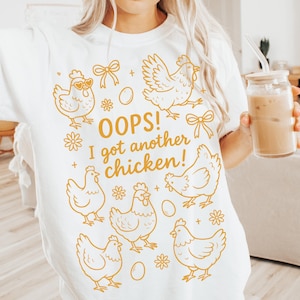 Könnte beinhalten: Weißes T-Shirt mit einer goldenen Illustration von Hühnern, Eiern, Schleifen und Blumen. Der Text "OOPS! I got another chicken!" ist in Gold gedruckt. Das Shirt ist ein lässiger, bequemer Stil.