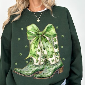 Puede incluir: Sudadera verde oscuro con un diseño de acuarela de botas de vaquero verdes decoradas con tréboles y un gran lazo verde. Las botas tienen un fondo blanco. Pequeños tréboles están esparcidos alrededor del diseño. Tema del Día de San Patricio.
