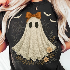 May include: Black t-shirt featuring a ghost design. The ghost is adorned with a bow and a lace-like sheet, surrounded by bats, spiderwebs, and floral elements. The design is in shades of cream, brown, and black.