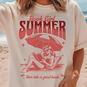 May include: A white t-shirt with a pink graphic of a woman reading a book under an umbrella on the beach. The text on the shirt reads "Book Girl Summer" and "Dive into a good book."
