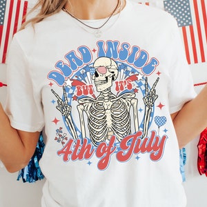 May include: White t-shirt with a skeleton wearing sunglasses and a red, white, and blue bandana. The skeleton is holding up peace signs and the text "Dead Inside But It's 4th of July" is printed on the shirt.