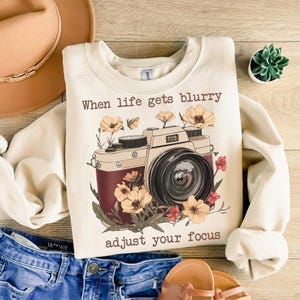 May include: A cream-colored sweatshirt with a vintage camera graphic and the text "When life gets blurry adjust your focus". The camera is surrounded by flowers in shades of red, yellow, and brown.