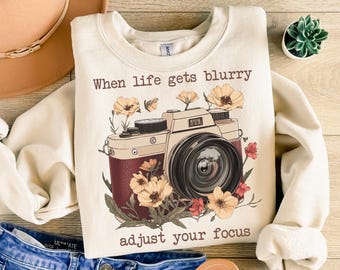 May include: A cream-colored sweatshirt with a vintage camera graphic and the text "When life gets blurry adjust your focus". The camera is surrounded by flowers in shades of red, yellow, and brown.