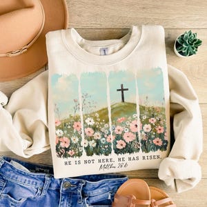 May include: A cream-colored sweatshirt with a graphic print of a cross on a hill with flowers. The text "HE IS NOT HERE. HE HAS RISEN. Matthew 28:6" is printed below the graphic.