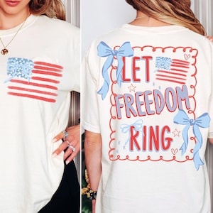 May include: Cream-colored t-shirt with a pocket featuring a painted American flag design. The back of the shirt has the phrase "LET FREEDOM RING" in red and blue, with bow and star accents. A patriotic themed tee.