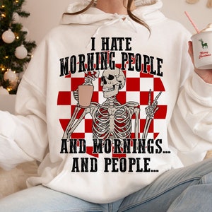 Puede incluir: Sudadera con capucha blanca con un gráfico de un esqueleto sosteniendo una taza de café y haciendo el signo de la paz. El texto dice: "I HATE MORNING PEOPLE AND MORNINGS... AND PEOPLE..." Un patrón de cuadros rojos y blancos está detrás del esqueleto.