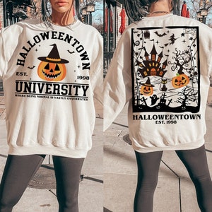 May include: Cream-coloured sweatshirt with "Halloweentown University" text and pumpkin graphic. The front design includes a pumpkin wearing a witch hat. The back features a Halloween-themed graphic with a haunted house, pumpkins, and bats. The text "EST. 1998" is included.