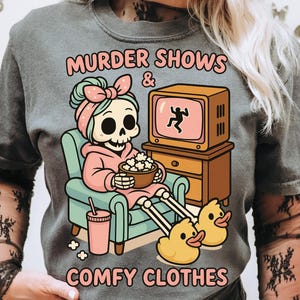 Op de afbeelding: Grijs T-shirt met de tekst "MURDER SHOWS & COMFY CLOTHES". De afbeelding toont een skelet in een roze badjas en eendenslippers, tv kijkend en popcorn etend. De tv toont een stokfiguur.