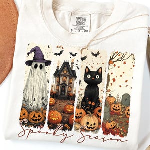 May include: Cream-colored t-shirt featuring a Halloween-themed design. The design includes a ghost wearing a witch hat, a haunted house, and a black cat surrounded by pumpkins. The words "Spooky Season" are written in a script font.