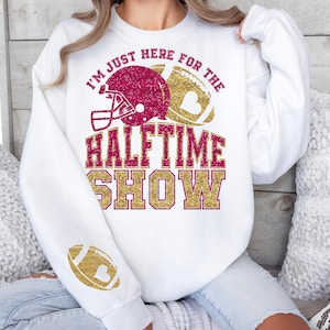 May include: White sweatshirt featuring a burgundy and gold glitter football helmet and football graphic. The text reads "I'M JUST HERE FOR THE HALFTIME SHOW." A gold football graphic is on the sleeve. A casual, comfortable top for game day.