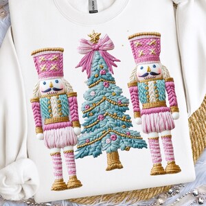 May include: White sweatshirt featuring a festive design with two nutcrackers and a Christmas tree. The nutcrackers have pink and blue accents, and the tree is adorned with gold ornaments. The design is embroidered on the front of the sweatshirt.