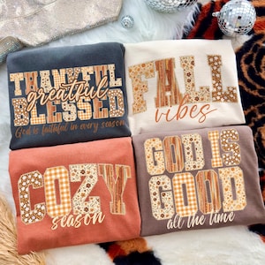 May include: Four sweatshirts with fall-themed text. The sweatshirts are in various colors: dark gray, cream, rust, and brown. The text includes "Thankful, Grateful, Blessed", "Fall Vibes", "Cozy Season", and "God is Good". The text is in a mix of floral and plaid patterns.