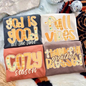 May include: Four folded sweatshirts in various colors, each featuring text in a patchwork style. The sweatshirts are in shades of gray, beige, coral, and brown. The text includes phrases like "God is Good," "Fall Vibes," "Cozy Season," and "Thankful, Grateful, Blessed."