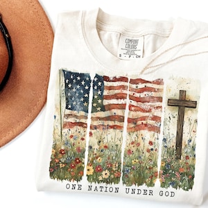 May include: A beige t-shirt with a watercolor design of an American flag with a cross in front of it. The flag is surrounded by flowers and the text "ONE NATION UNDER GOD" is printed below the design.