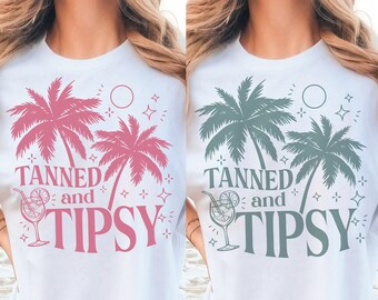 May include: Two white t-shirts with a graphic design of palm trees, a cocktail, and the text "Tanned and Tipsy". One shirt has pink graphics and the other has green graphics.