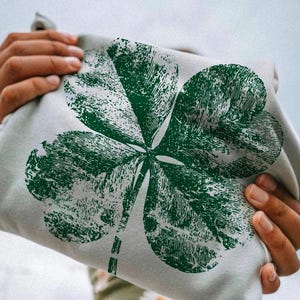 Könnte beinhalten: Hellgraues Sweatshirt mit einem großen, verwaschenen grünen vierblättrigen Kleeblatt-Design. Das Kleeblatt ist mittig auf der Vorderseite. Das Sweatshirt wird von einer Person hochgehalten.