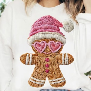 Op de afbeelding: Witte sweatshirt met een geborduurd peperkoekenmannetje. Het peperkoekenmannetje draagt een roze kerstmuts en een hartvormige bril. Het heeft bruine en witte details, met roze knopen. Een feestelijk en grillig ontwerp.