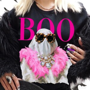May include: A black long-sleeve shirt featuring a graphic of a ghost wearing leopard print sunglasses and a pink feather boa with a large crystal necklace. The word BOO is printed in large hot pink letters above the ghost. The shirt is worn under a black faux fur jacket.
