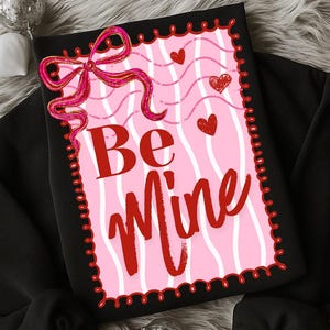 May include: Black sweatshirt with a pink and white striped graphic that says "Be Mine" in red lettering. The graphic includes red hearts and a pink bow. The sweatshirt is displayed on a fluffy, gray surface.