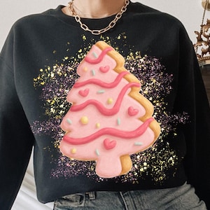 Puede incluir: Sudadera negra con un diseño de árbol de Navidad rosa y amarillo con detalles de corazones rosas. El gráfico está rodeado de un efecto de salpicaduras coloridas. La sudadera se combina con vaqueros rotos de color azul claro.