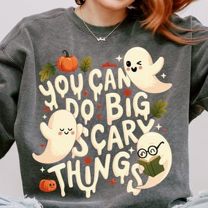 Könnte beinhalten: Dunkelgraues Sweatshirt mit dem Schriftzug "You Can Do Big Scary Things" und Illustrationen von Geistern, Kürbissen und Blättern. Das Design ist in Weiß, Orange und Grün gehalten. Ein lässiges Kleidungsstück zum Thema Herbst.