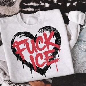 May include: A light gray sweatshirt with a black heart design and the words "FUCK ICE" in red. The design has a distressed, grunge style with dripping paint effects. The sweatshirt is paired with denim and a checkered shoe.