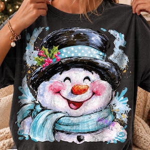 May include: A charcoal gray short-sleeve t-shirt featuring a graphic of a smiling snowman wearing a black top hat with a light blue polka dot band, a light blue scarf, and a sprig of holly with red berries, set against a distressed blue background.