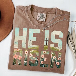 May include: A brown t-shirt with a faded look featuring the words "He is Risen" in large, block letters. The letters are filled with a floral design and a cross is visible in the background. The text "In Matthew 28:6" is printed in smaller letters below the main text.