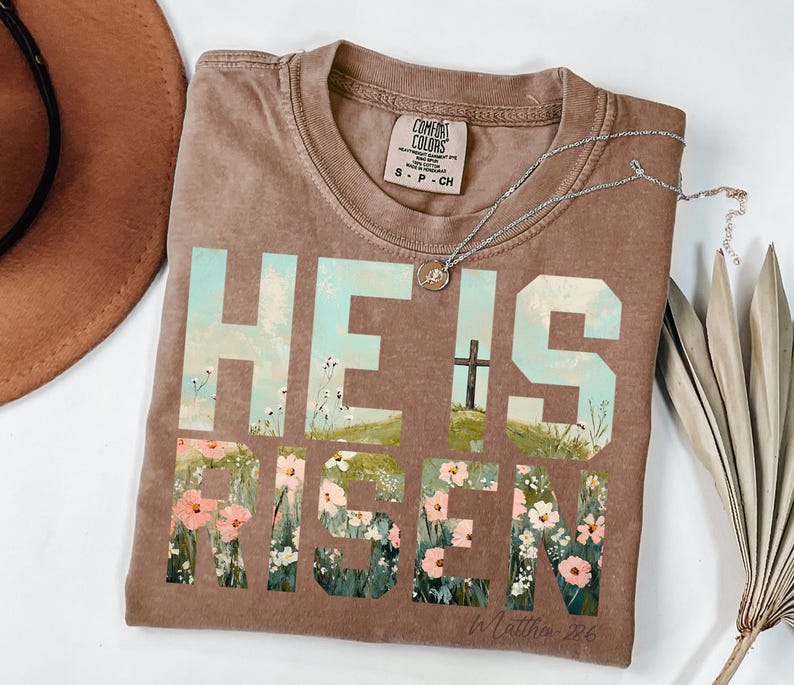 May include: A brown t-shirt with a faded look featuring the words "He is Risen" in large, block letters. The letters are filled with a floral design and a cross is visible in the background. The text "In Matthew 28:6" is printed in smaller letters below the main text.