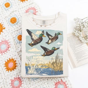 May include: Cream-colored t-shirt featuring a vintage-style illustration of three ducks in flight over water and reeds. A delicate necklace and a book with flowers are next to the shirt. The shirt has the text "Comfort Classic".