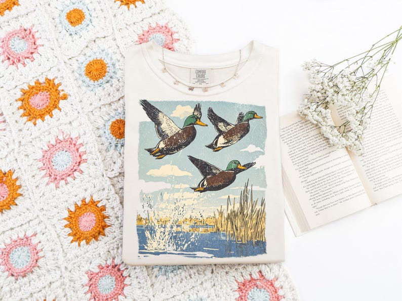May include: Cream-colored t-shirt featuring a vintage-style illustration of three ducks in flight over water and reeds. A delicate necklace and a book with flowers are next to the shirt. The shirt has the text "Comfort Classic".