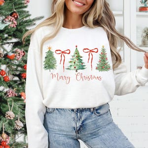 May include: White crew neck sweatshirt with a watercolor design of four Christmas trees and red bows. The words "Merry Christmas" are written in red script below the trees. The sweatshirt is paired with blue jeans.