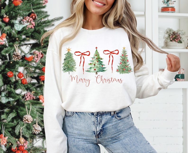 May include: White crew neck sweatshirt with a watercolor design of four Christmas trees and red bows. The words "Merry Christmas" are written in red script below the trees. The sweatshirt is paired with blue jeans.