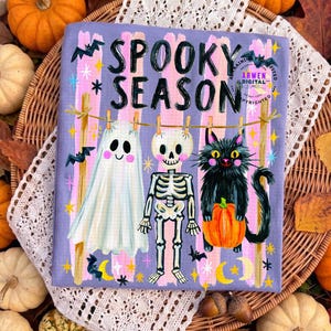 K&ouml;nnte beinhalten: Ein rechteckiges Stoffpaneel mit dem Text SPOOKY SEASON in schwarzen Buchstaben. Das Design zeigt ein wei&szlig;es Gespenst, ein Skelett und eine schwarze Katze mit einem orangefarbenen K&uuml;rbis, die an einer W&auml;scheleine h&auml;ngen. Der Hintergrund hat vertikale rosa und violette Streifen mit kleinen gelben Sternen, Flederm&auml;usen und Mondsicheln.