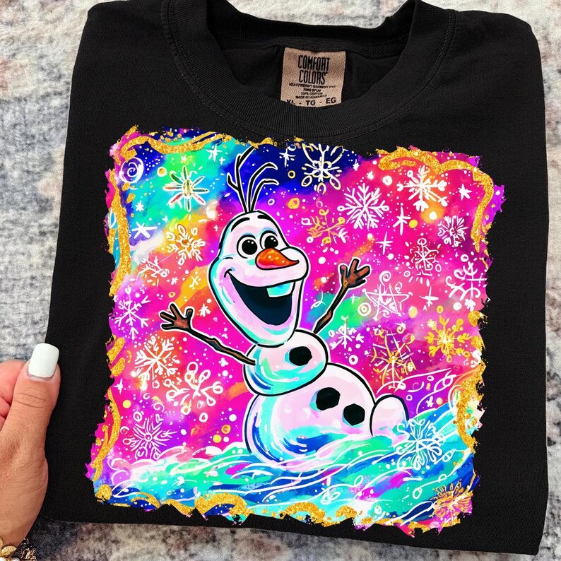以下が含まれることがあります： 黒色の長袖Tシャツ。アナと雪の女王の雪だるま、オラフのカラフルなグラフィックが特徴です。デザインには雪の結晶と、ピンク、青、金色の鮮やかな背景が含まれています。Tシャツはクルーネックです。