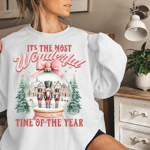 May include: White sweatshirt with a pink and red graphic design. The graphic features a snow globe with three nutcracker figurines inside. The text on the sweatshirt reads "It's the most Wonderful Time of the Year."