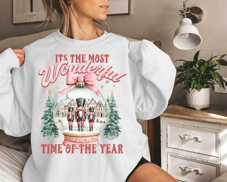 May include: White sweatshirt with a pink and red graphic design. The graphic features a snow globe with three nutcracker figurines inside. The text on the sweatshirt reads "It's the most Wonderful Time of the Year."