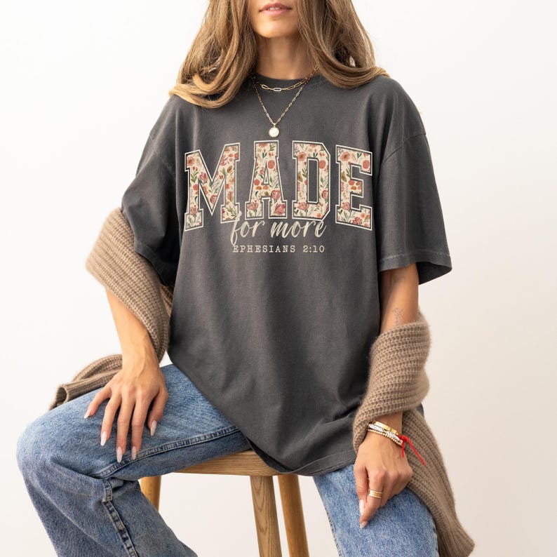 Op de afbeelding: Donkergrijs T-shirt met de tekst "MADE for more" in een bloemenpatroon. De tekst "Ephesians 2:10" staat eronder. Het shirt wordt gecombineerd met een blauwe jeans en een beige vest.