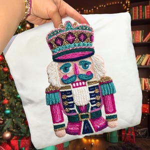 May include: A white pillow featuring a colorful, beaded nutcracker design. The nutcracker has a pink and teal uniform, a gold crown, and a white beard. The design is intricately detailed, with various bead colors and textures, creating a festive holiday theme.