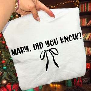 Może przedstawiać: Biała poszewka na poduszkę z czarnym napisem "MARY, DID YOU KNOW?" oraz czarną grafiką kokardy. Poszewka jest trzymana przez rękę, w tle choinka i regał z książkami.