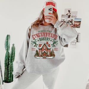 May include: A light gray sweatshirt and sweatpants set. The sweatshirt features a graphic of a gingerbread house with the text "North Pole Gingerbread Bakery". A red and white hat is worn.