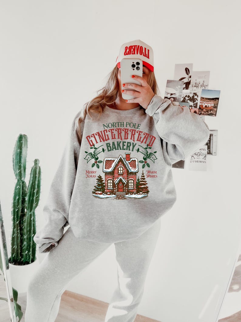 May include: A light gray sweatshirt and sweatpants set. The sweatshirt features a graphic of a gingerbread house with the text "North Pole Gingerbread Bakery". A red and white hat is worn.