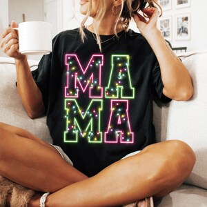 May include: Black t-shirt with the word "MAMA" in large, colorful, neon-style letters. The letters are outlined in green and pink, filled with small, colorful lights. The shirt is worn by a person sitting on a couch.