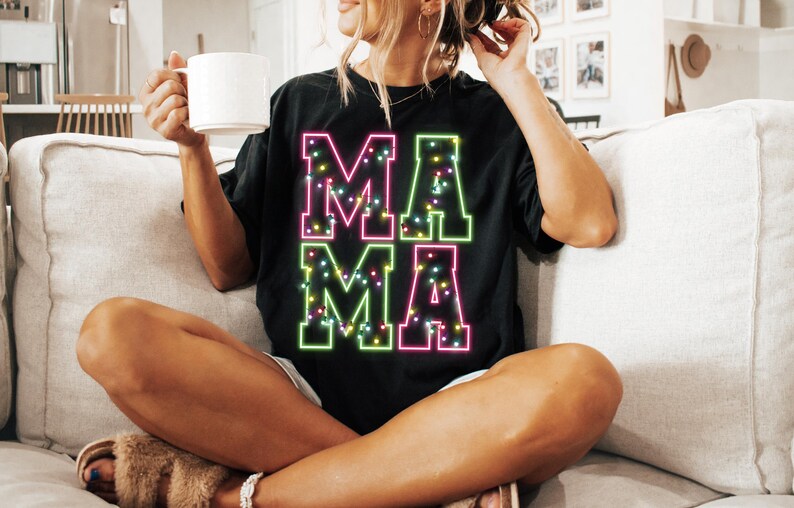 May include: Black t-shirt with the word "MAMA" in large, colorful, neon-style letters. The letters are outlined in green and pink, filled with small, colorful lights. The shirt is worn by a person sitting on a couch.