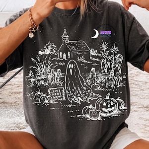 May include: A charcoal gray t-shirt featuring a white line art graphic of a haunted scene with a church, a ghost, corn stalks, and pumpkins. A sign in the graphic reads "LIVE HERE NOW". The design includes a crescent moon and circular text.