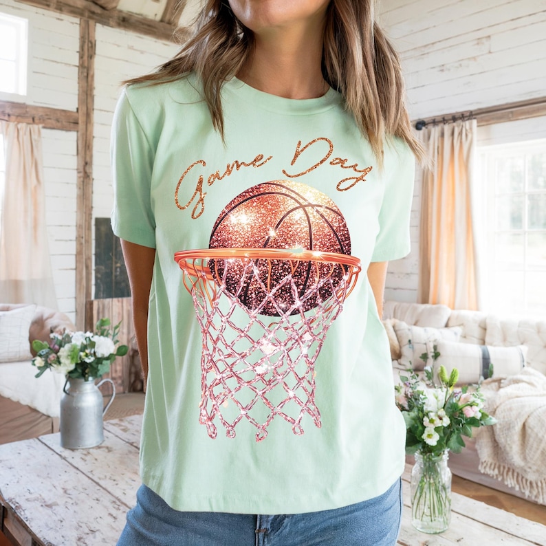 May include: A light green t-shirt with the text "Game Day" in gold glitter lettering. A glittery gold basketball is shown going through a pink glitter basketball hoop.