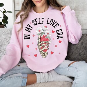 May include: A light pink sweatshirt with the text "IN MY SELF LOVE ERA" in black. The graphic features a skeletal hand holding a red heart with white daisies. Red hearts and stars are scattered around the graphic.