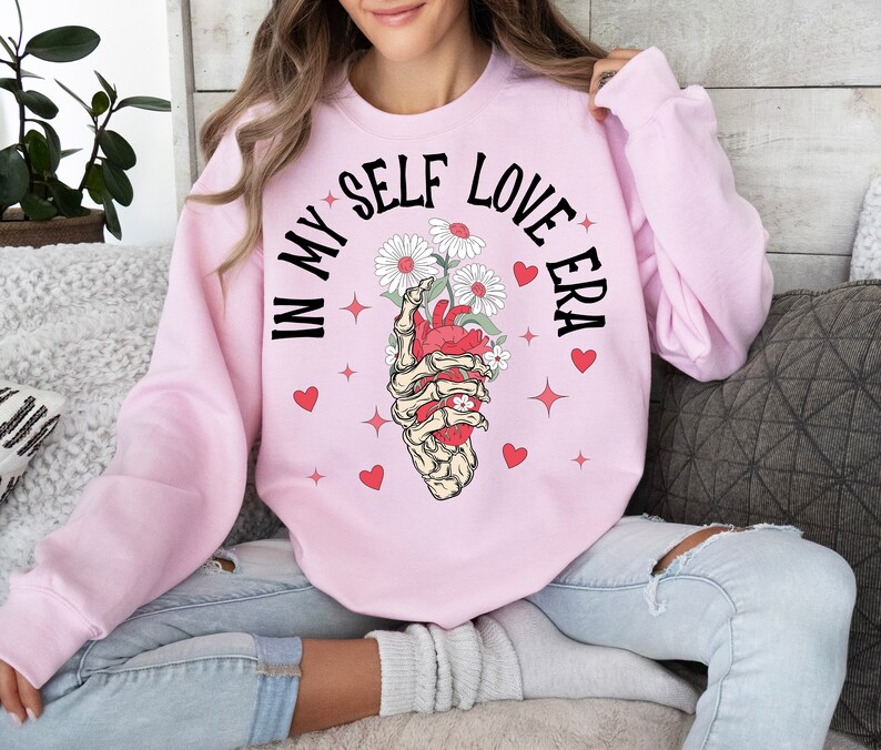 May include: A light pink sweatshirt with the text "IN MY SELF LOVE ERA" in black. The graphic features a skeletal hand holding a red heart with white daisies. Red hearts and stars are scattered around the graphic.