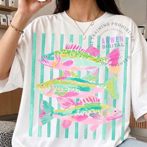 May include: White t-shirt featuring a colorful fish design. The design includes three fish in shades of pink, green, and yellow, set against a backdrop of vertical teal stripes. The text "ARWEN DIGITAL" is visible.
